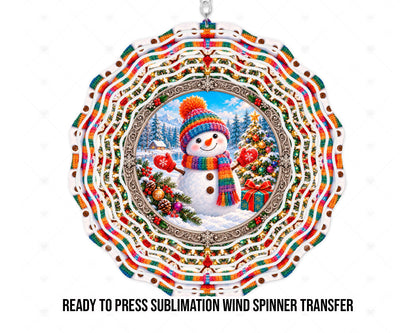 Wind spinner mockup showing snowman artwork for a physical printed sublimation transfer. A smiling snowman in a rainbow knit hat and scarf wears red snowflake mittens beside a decorated Christmas tree and gift. Snowy evergreens, mountains, and a cabin sit behind an ornate silver-toned frame, with rainbow stripes and holiday motifs across concentric scalloped rings.