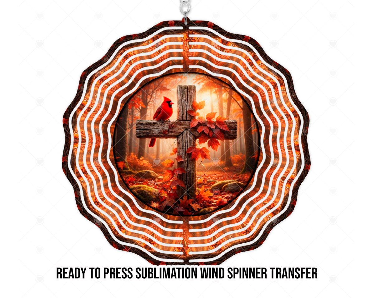 Mockup of a wind spinner sublimation transfer design featuring a red cardinal perched on a rustic wooden cross entwined with red and orange leaves. A sunlit autumn forest, fallen leaves, and mossy rocks fill the background. Concentric scalloped rings carry the warm woodland colors around the central artwork.